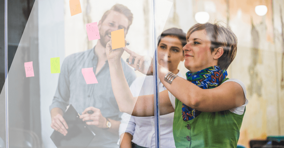 Photo de trois personnes travaillant ensemble dans une séance de brainstorming, utilisant des notes adhésives colorées sur une vitre. Une femme au premier plan écrit sur la vitre pendant que les autres observent.